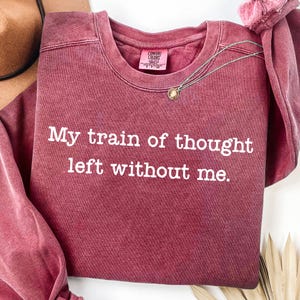 May include: A maroon crewneck sweatshirt with a vintage-washed finish featuring the white text My train of thought left without me. The garment includes a Comfort Colors brand label inside the collar and a thin silver chain necklace resting on top.