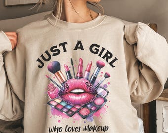 May include: Beige sweatshirt with the text "JUST A GIRL" above a graphic of pink lips, makeup brushes, lipstick, and an eyeshadow palette. The text "who loves Makeup" is below the graphic. The design has a watercolor effect.
