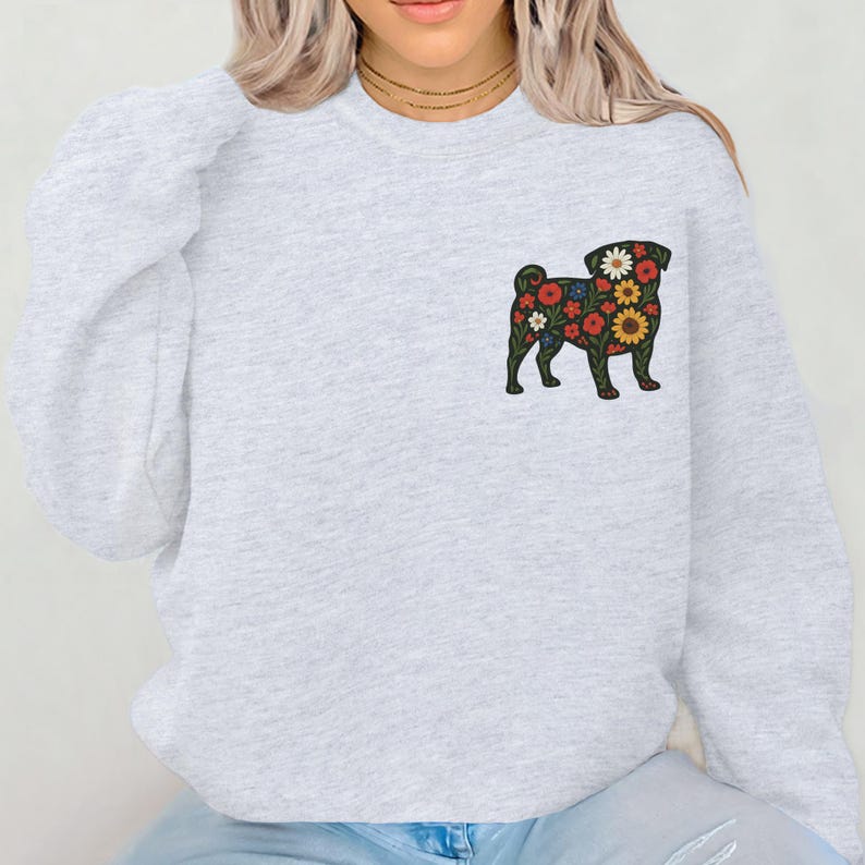 May include: A light gray sweatshirt featuring a floral pug design on the left chest. The pug is black with a colorful floral pattern of red, yellow, and white flowers. The sweatshirt has a crew neck and long sleeves.