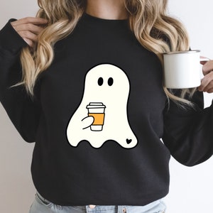 May include: Black sweatshirt with a white ghost holding a cup of coffee. The ghost has two black eyes and a small heart on its chest.