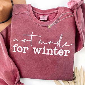 May include: A maroon crewneck sweatshirt with the text "not made for winter" in white script. A gold pendant necklace and a brown hat are visible. The sweatshirt has a vintage, washed look.