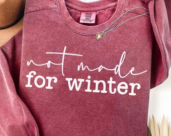 Puede incluir: Una sudadera burdeos con la frase "not made for winter" en escritura blanca. Se ve un collar con colgante dorado y un sombrero marrón. La sudadera tiene un aspecto vintage y lavado.