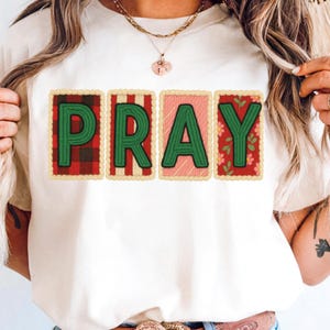 May include: Cream-colored t-shirt featuring the word "PRAY" in green letters. Each letter is set against a different patterned background: plaid, stripes, pink, and floral. The design has a stitched, patch-like appearance.