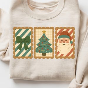 Peut inclure: Sweat-shirt crème avec trois motifs rectangulaires sur le thème de Noël. Les motifs comprennent un nœud vert, un sapin de Noël avec des boules rouges et le Père Noël. Les motifs sont encadrés d'un bord festonné.