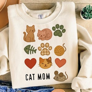 Puede incluir: Sudadera de color crema con varios apliques con temática de gatos. Los diseños incluyen gatos, huellas de patas, corazones, un esqueleto de pez, una bola de hilo y un ratón. Las palabras "CAT MOM" están impresas en letras negras en la parte inferior.