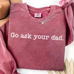 Puede incluir: Una sudadera Comfort Colors color granate con la frase "Go ask your dad" en letras blancas. Un collar plateado con un colgante está sobre el frente. Un sombrero marrón es visible en el fondo. La sudadera está doblada.