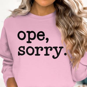 Peut inclure: Un sweat-shirt rose avec le texte "ope, sorry." imprimé en noir.