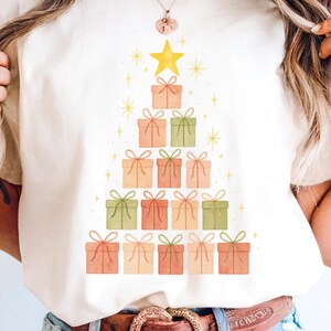 Pode incluir: Camiseta branca com um design de árvore de Natal em aquarela. A árvore é composta por caixas de presente em tons de pêssego, verde e coral, encimada por uma estrela amarela. Explosões de estrelas douradas cercam a árvore, criando um tema festivo.