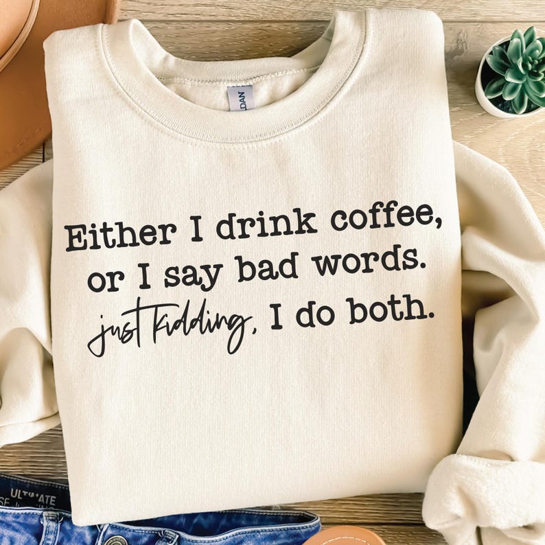 May include: Cream-colored sweatshirt with the text "Either I drink coffee, or I say bad words. Just kidding. I do both." in black lettering. The sweatshirt is folded and displayed on a wooden surface.