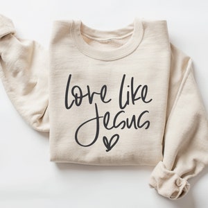 May include: A beige sweatshirt with the text "love like Jesus" in black script with a small heart below the text.