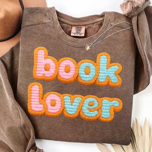 Könnte beinhalten: Ein braunes Sweatshirt mit den Worten "book lover" in Rosa und Blau mit orangefarbenen Konturen gestickt. Das Sweatshirt hat einen Rundhalsausschnitt und lange Ärmel. Eine silberne Halskette mit einem kleinen Anhänger ist sichtbar.
