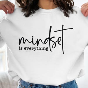 Puede incluir: Sudadera blanca de manga larga con la frase "mindset is everything" en escritura negra. El texto está centrado en la parte delantera. La sudadera está hecha de un material suave.