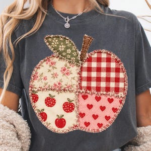 May include: A charcoal gray t-shirt featuring a large, central apple-shaped applique. The apple is divided into four sections with different fabric patterns: floral, red and white gingham, small red apples on cream, and red hearts on pink.