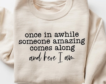 May include: A beige sweatshirt with the text "once in awhile someone amazing comes along and here I am" printed in black.