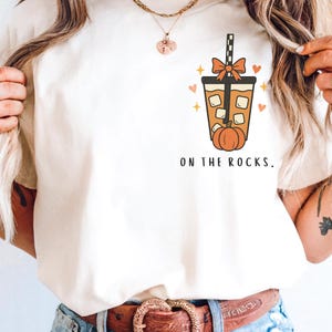 以下が含まれることがあります： クリーム色のTシャツ。秋をテーマにしたグラフィックが特徴です。デザインには、氷、リボン、カボチャが入ったパンプキン スパイス ドリンクが含まれています。「ON THE ROCKS」の文字が下にプリントされています。カジュアルな季節の衣料品です。