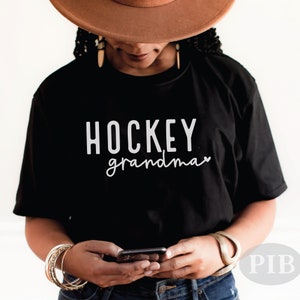 May include: A black t-shirt with the text "HOCKEY grandma" in white lettering. The shirt is being worn by a person with brown skin and brown hair. The person is wearing a brown hat and holding a black smartphone.