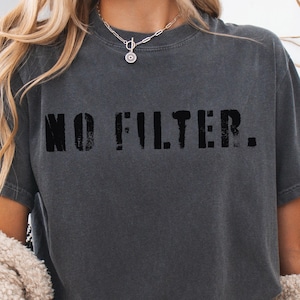 Puede incluir: Camiseta gris oscuro con la frase "NO FILTER." impresa en letras negras desgastadas. La camiseta tiene mangas cortas y cuello redondo. La tela parece suave y ligeramente descolorida.