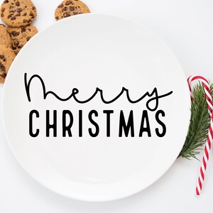 May include: White ceramic plate with the text "Merry Christmas" in black lettering. The plate is surrounded by cookies and a candy cane.