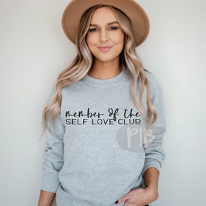 May include: A gray sweatshirt with the text "Member of the Self Love Club" in black lettering. The sweatshirt has a round logo with the letters "PLB" in black.