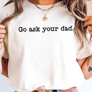 May include: Cream-colored t-shirt with the phrase "Go ask your dad" printed in black. The shirt is a casual style, suitable for everyday wear. The text is centered and in a bold, easy-to-read font.