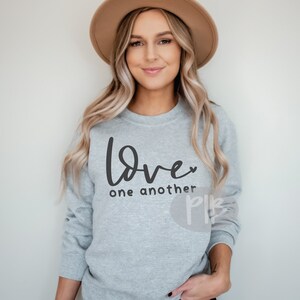 May include: A light gray sweatshirt with the words "Love one another" in black script. The word "Love" has a small heart replacing the "o". The sweatshirt is long-sleeved and has a crew neck.