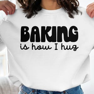 May include: White sweatshirt with black text that reads "Baking is how I hug."