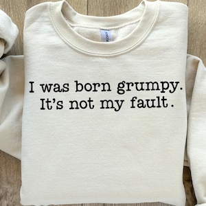 May include: A cream-colored sweatshirt with the text "I was born grumpy. It's not my fault." in black. The sweatshirt has a crew neck and long sleeves, and is folded neatly.