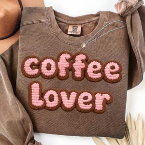 May include: A brown sweatshirt with the words "coffee Lover" in a pink and brown textured design. A silver necklace with a small pendant is draped on the shirt. The shirt has a crew neck and short sleeves.