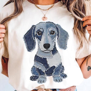 May include: A cream-coloured t-shirt with a denim patchwork dachshund dog design. The dog is made from various shades of blue denim, with a black nose and large eyes. The design is centrally placed on the shirt.