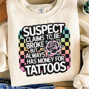 May include: A cream coloured sweatshirt with a colourful graphic that says "SUSPECT CLAIMS TO BE BROKE BUT ALWAYS HAS MONEY FOR TATTOOS". The graphic is in a checkered pattern with a pink rose.