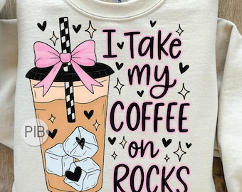 May include: A cream-coloured sweatshirt with a graphic of an iced coffee, pink bow, black straw, and ice cubes. The text "I take my coffee on rocks" is in black and pink, with hearts and stars. A casual top.