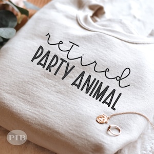 May include: A white sweatshirt with the text "retired party animal" printed in black. The sweatshirt is folded and a gold necklace is lying on top of it.
