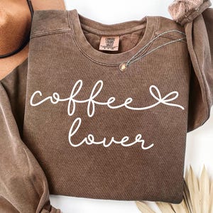 May include: A brown crewneck sweatshirt with the words "coffee lover" in white script. A silver necklace with a small pendant rests on the shirt. The sweatshirt has a vintage, washed look.