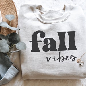 Może przedstawiać: Biała bluza z nadrukiem "fall vibes" w kolorze czarnym. Tekst jest w retro stylu czcionki.