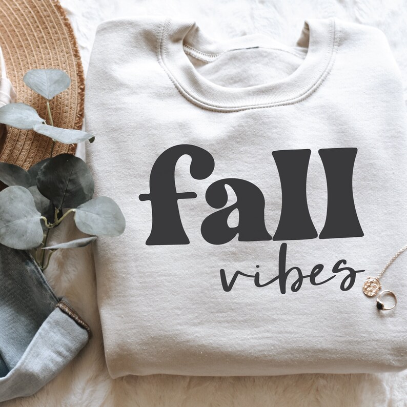 Może przedstawiać: Biała bluza z nadrukiem "fall vibes" w kolorze czarnym. Tekst jest w retro stylu czcionki.