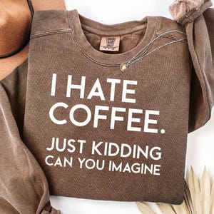 Puede incluir: Una sudadera marrón Comfort Colors con el texto "I HATE COFFEE. JUST KIDDING CAN YOU IMAGINE" en blanco. Un collar plateado con un colgante está drapeado sobre la camisa. Un sombrero marrón es visible en el fondo.