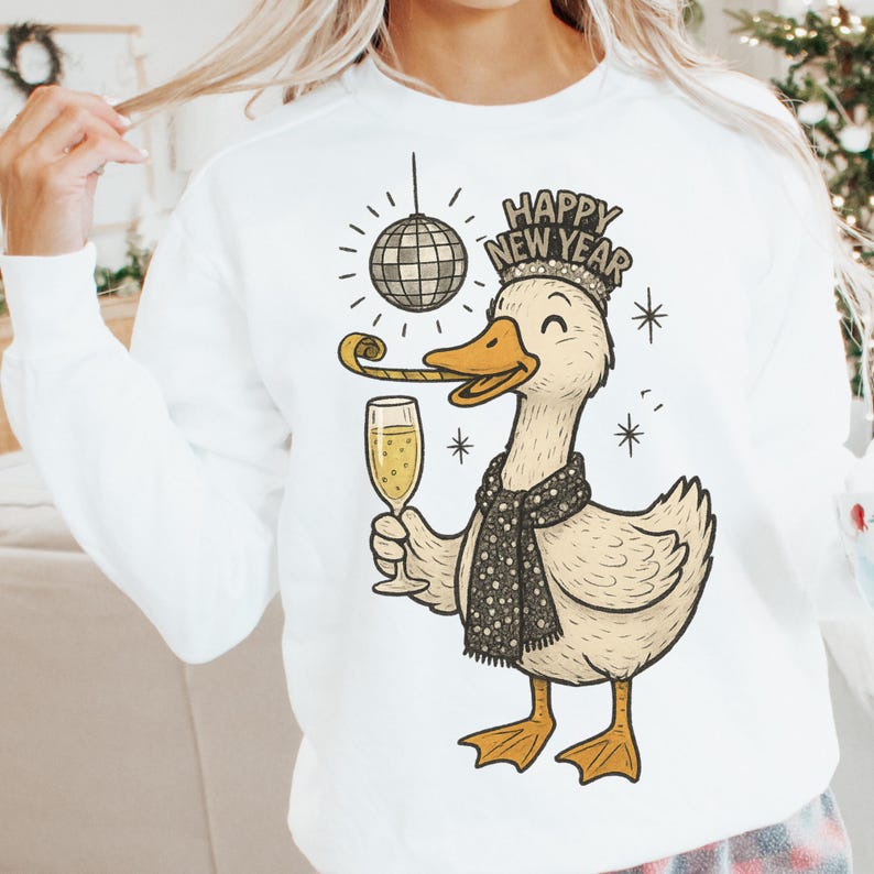 May include: White sweatshirt featuring a cartoon duck celebrating New Year's. The duck wears a Happy New Year crown and a scarf, holding a champagne flute and party horn. A disco ball hangs above the duck.