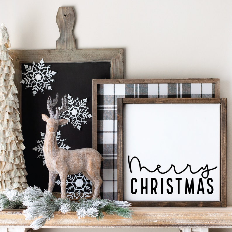May include: A wooden sign with the text "Merry Christmas" in black script font. The sign is framed in a dark wood frame and is sitting on a shelf with a small, brown, deer figurine and a white, paper Christmas tree.