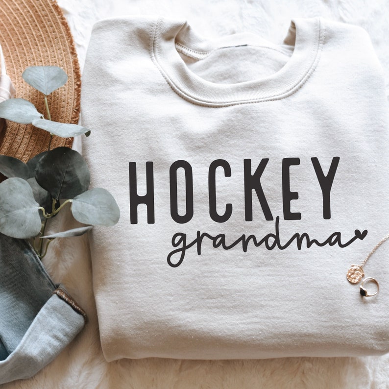 May include: A light grey sweatshirt with the text "HOCKEY grandma" in black lettering. The text is in a casual font with a heart symbol after the word "grandma".