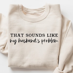 Może przedstawiać: Kremowa bluza z czarnym napisem "THAT SOUNDS LIKE my husband's problem". Bluza ma okrągły dekolt i długie rękawy. Tekst jest pogrubiony, czcionką bezszeryfową.