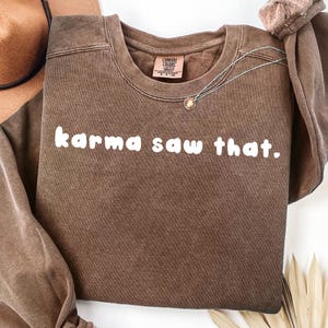 May include: A brown crewneck sweatshirt with the phrase "karma saw that" in white block letters. A silver necklace with a pendant is draped across the front. The sweatshirt has a vintage, washed look.