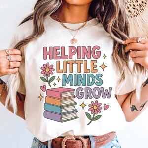 May include: Cream-colored t-shirt with the text "HELPING LITTLE MINDS GROW" in colorful, retro-style lettering. The design includes a stack of books, flowers, hearts, and stars, perfect for teachers or educators.