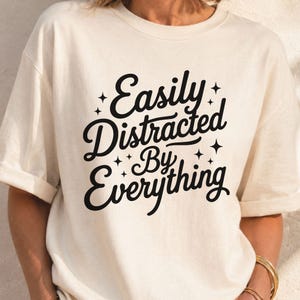 Può includere: Una t-shirt a maniche corte color crema con la scritta nera "Easily Distracted By Everything" in un carattere corsivo stilizzato, accentuata da piccole icone a forma di stella a quattro punte sparse attorno alla scritta sul davanti.