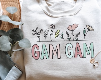 May include: A cream-coloured sweatshirt featuring the words "GAM GAM" in pastel shades, with a floral design above. The flowers are in pink, yellow, and green. The sweatshirt is folded on a white surface, with a straw hat visible.