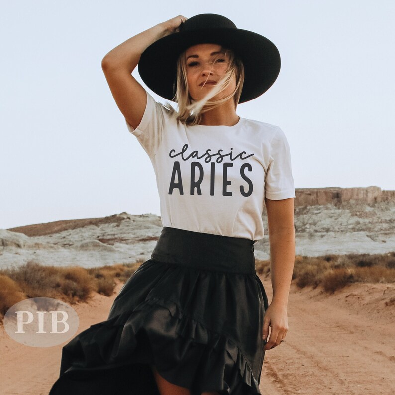 Puede incluir: Una camiseta blanca con el texto "classic ARIES" en letras negras. La persona que lleva la camiseta est&aacute; de pie en un paisaje des&eacute;rtico y lleva un sombrero negro y una falda negra.