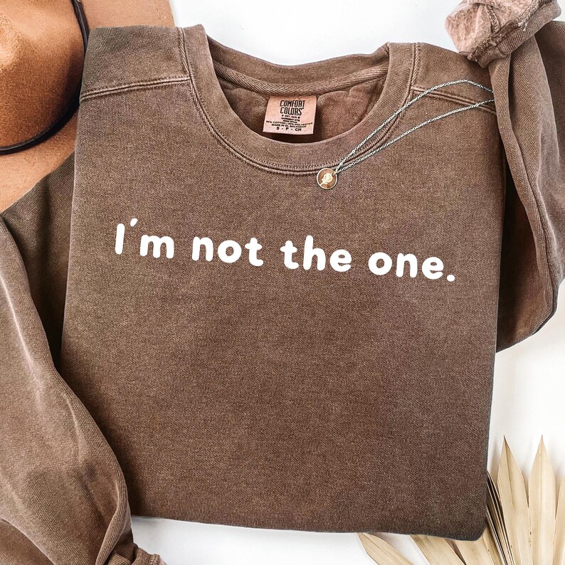May include: A brown crewneck sweatshirt with the phrase "I'm not the one" printed in white. A silver necklace with a pendant is draped on the shirt. The sweatshirt has a vintage, washed look.
