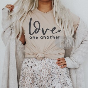 May include: Beige t-shirt with the words "Love one another" in dark gray script. The shirt is knotted at the waist and worn with a patterned skirt and a light gray cardigan. The overall style is casual and comfortable.
