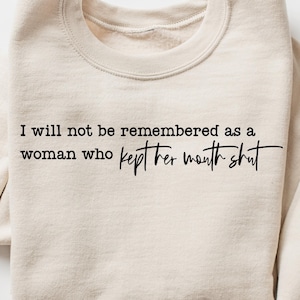 May include: A cream-coloured sweatshirt featuring the quote, "I will not be remembered as a woman who kept her mouth shut" in black text. The sweatshirt has a crew neck and long sleeves.