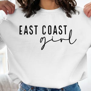 Puede incluir: Sudadera blanca con el texto "East Coast girl" en letras negras.