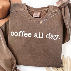 May include: A brown Comfort Colors sweatshirt with the phrase "coffee all day" in white block letters. A silver necklace with a small pendant is draped across the front. The sweatshirt is folded, with a brown hat visible in the background.